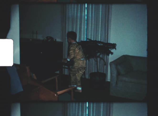 A small boy wearing yellow and brown tiger pajamas hides toys across a dimly lit living room with dark furniture