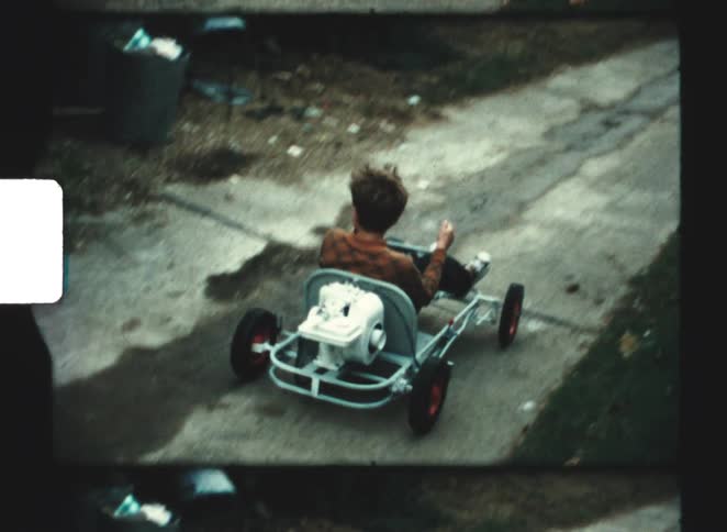 A boy in a plaid shirt is sitting in a small homemade go kart with a white engine and drives away down a narrow paved driveway