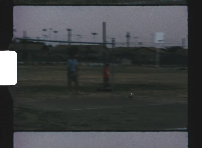 A young boy in a white shirt plays soccer on a grassy sports field near a goalpost in vintage film footage