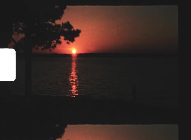 An orange sun sits low on the horizon casting a vertical reflection across calm water with a dark tree silhouette on the left a distant shoreline in the background and a person standing near the waters edge in shadow