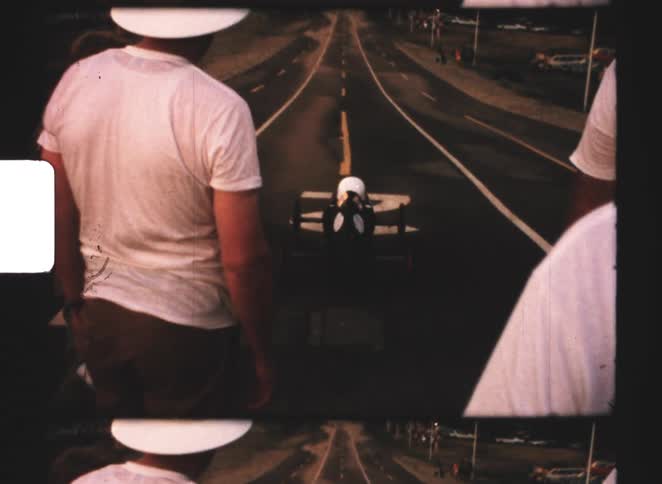 From behind people in white shirts and sun hats observe a small blackandwhite race car or gokart positioned on a paved track marked with a large 2