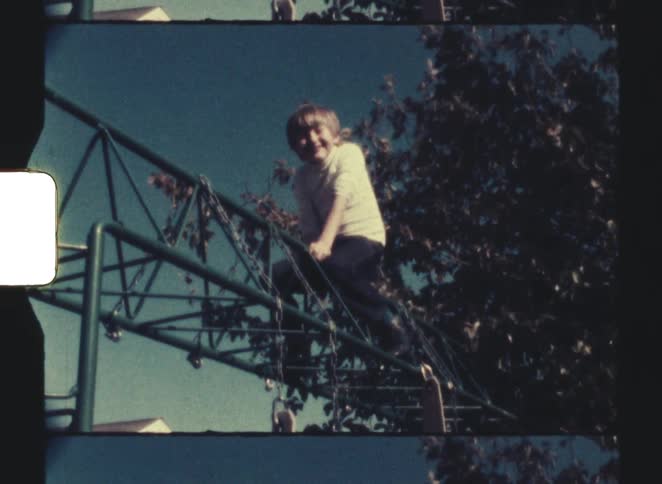 A young boy with short light hair is seen smiling and clinging to the top bar of a green metal jungle gym against a backdrop of dark trees and a clear blue sky