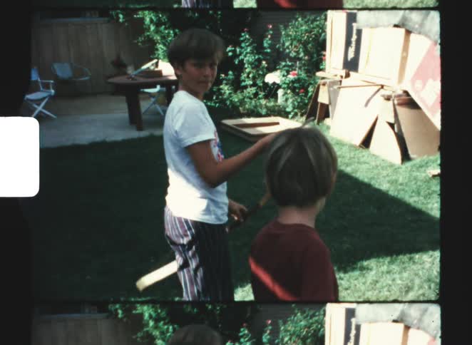 A boy in a white shirt plays with a makeshift bow and arrow decorated with colorful ribbons in a grassy backyard near a house