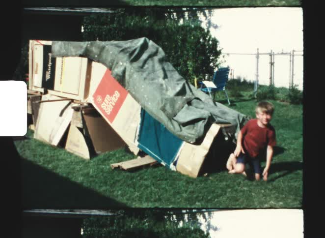 A young boy crawls out of an elaborate makeshift fort built from cardboard appliance boxes and a tarp standing up on the grassy lawn in this nostalgic home movie