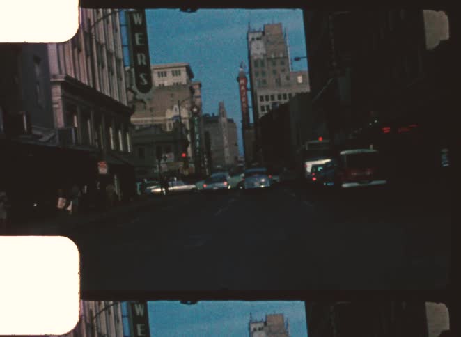 A vintage film frame shows a busy downtown city street with traffic and tall buildings under a muted blue sky