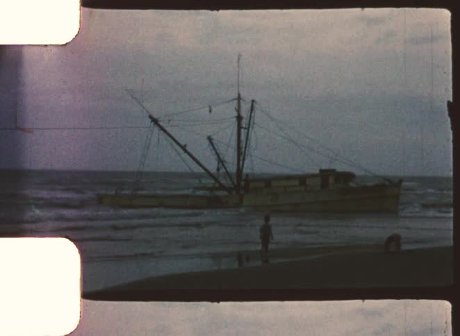 Two kids playing on the beach at sunset with a wrecked ship along the shoreline in the background and waves hitting the shore