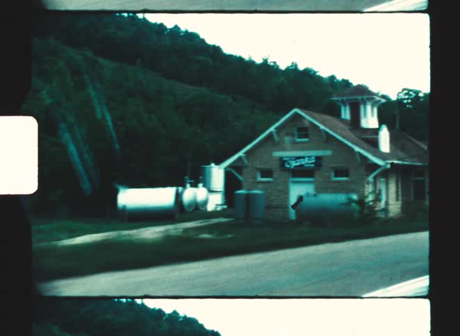 Brick building with pitched roof stands beside cylindrical tanks and dense vegetation