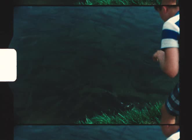 A young boy in a blue and white striped shirt stands on a grassy bank leaning over to throw an object into the dark water causing ripples to spread across the surface