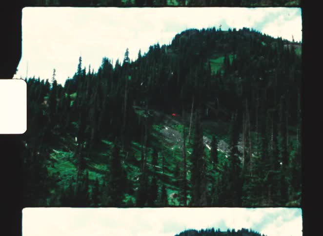 Mountain landscape covered in pine trees under partly cloudy sky framed by film borders