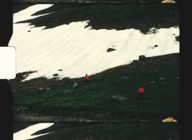 Three people in red jackets walking across mountainous terrain with snow and vegetation