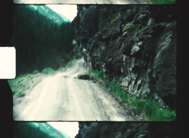 A blurry point of view shot travels down a narrow dirt road surrounded by steep rocky and forested cliffs in vintage film