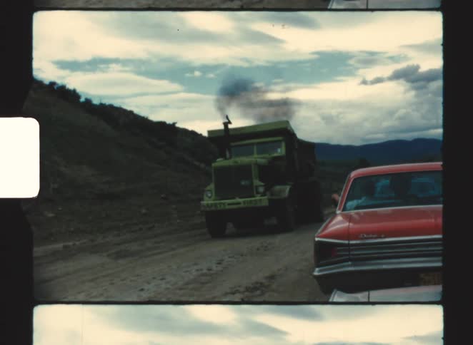 A trailer driving along an untarred road at midday passing an orange car parked on the roadside