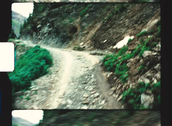 Narrow trail on a rocky hillside surrounded by vegetation with mountains in the background