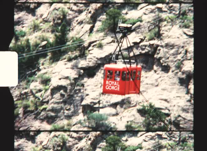 Vintage footage captures a bright red aerial cable car stamped with royal gorge drifting slowly past the rugged sunlit rock face of a steep canyon wall