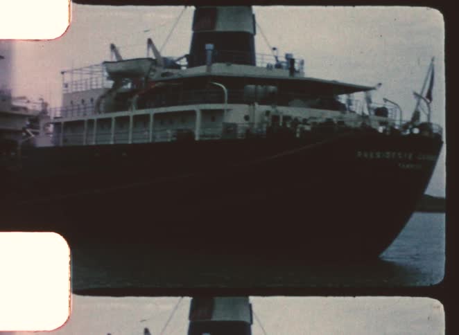 A static shot of a large dark cargo ship pulling away from a dock or being viewed from a structure on a dull overcast day