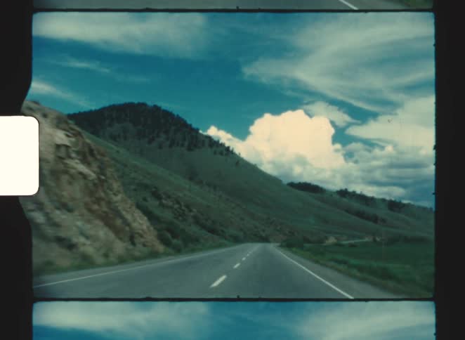 Twolane highway winding past grassy hills beneath blue sky with dense clouds