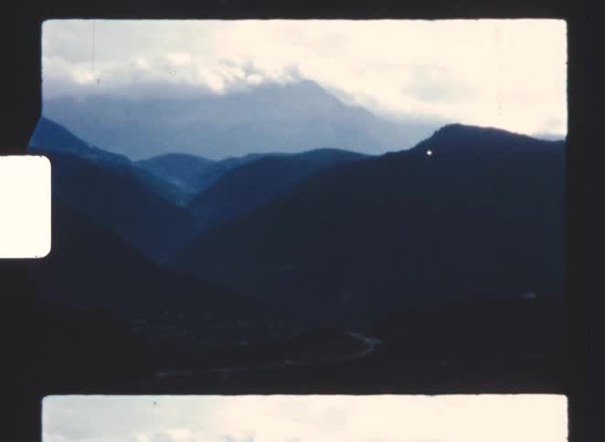 A vintage film view captures a deep valley surrounded by dark tiered mountain ranges under a cloudy sky