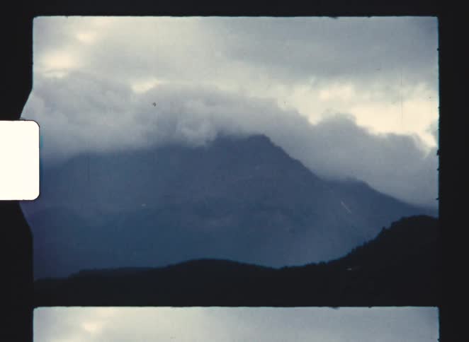 This is a very short video clip approximately 3 seconds that appears to be footage from an old film reel perhaps 8mm or 16mm indicated by the border and film sprocket hole visible on the left sidethe primary subject is a dark cloudshrouded mount