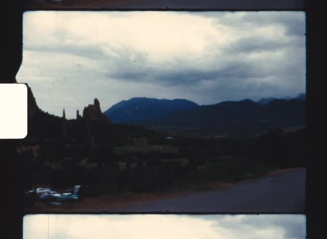 Curved road passes parked vehicles before vertical rock formation and mountains under cloudy sky