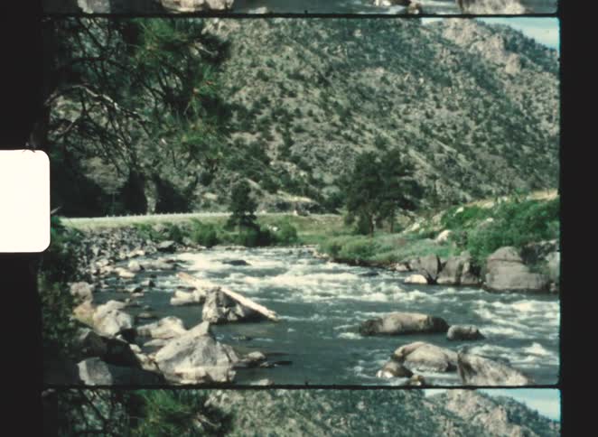 Car travels along a highway between a rapidly flowing stream and a mountain on a cloudy summer day