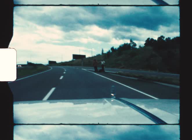 People standing by the roadside with luggage beside them and vegetation in the background