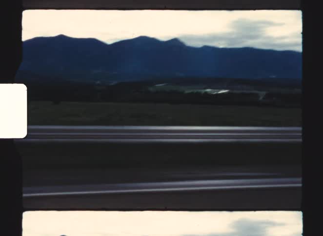 A blurred multilane road runs in the foreground with dark mountains and a field in the distance under an overcast sky