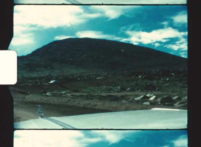 A car driving through the countryside at midday with hills and rocky terrain lining the side of the road