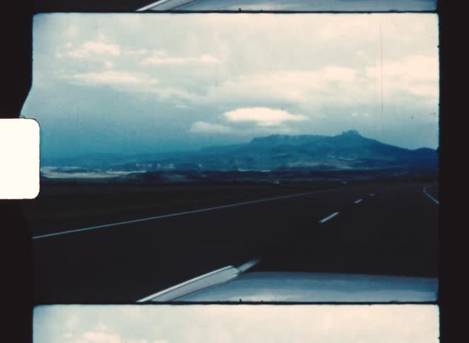 Captured from the passenger seat of a moving vehicle the camera looks out over the white hood as the car travels down a paved highway toward a towering blue mountain range shrouded in clouds