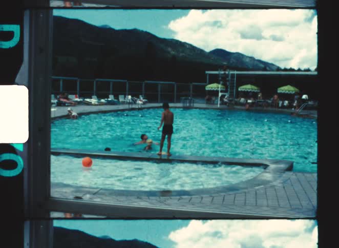 People relaxing around and swimming in an outdoor pool on a sunny day with a child standing at the edge of the shallow end while a large forested mountain range rises prominently in the background