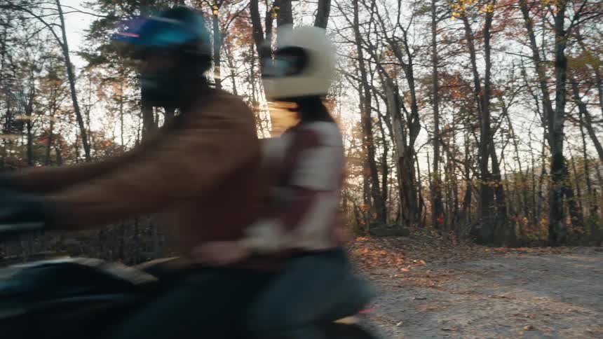 Two people wearing helmets ride a motorcycle along a road surrounded by trees
