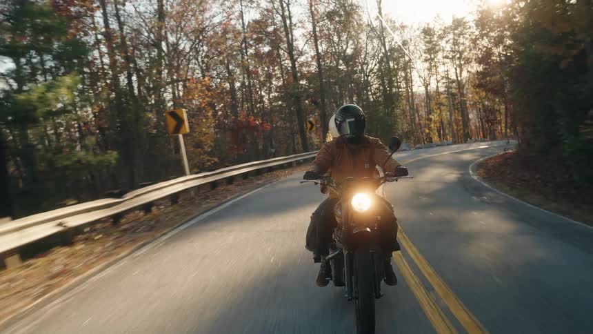 A person rides a motorcycle with its headlight on navigating a winding forest road as the setting sun creates a brilliant flare