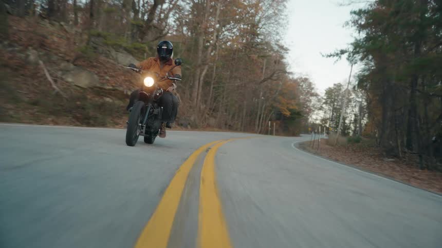 A motorcyclist wearing a tan shirt and black helmet cruises along a winding country road leaning smoothly into the curves amidst a backdrop of rustcolored autumn foliage