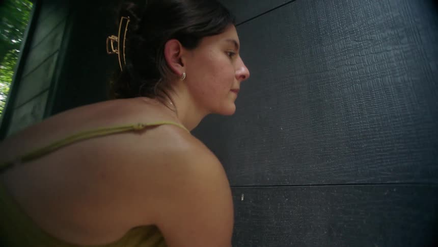 A woman with dark hair in a loose topknot uses her arm to open a dark wooden door to a ceramics studio in a forest setting