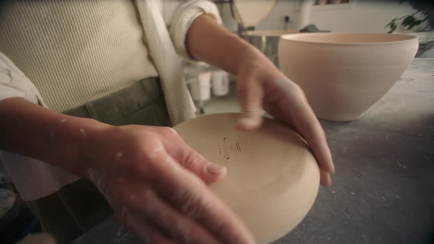 Hands smooth the base of an unfired ceramic piece on a workbench
