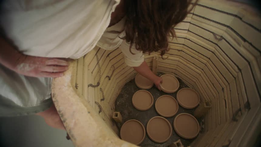 A woman places a freshly shaped clay bowl into an open pottery kiln already partially filled with other pieces