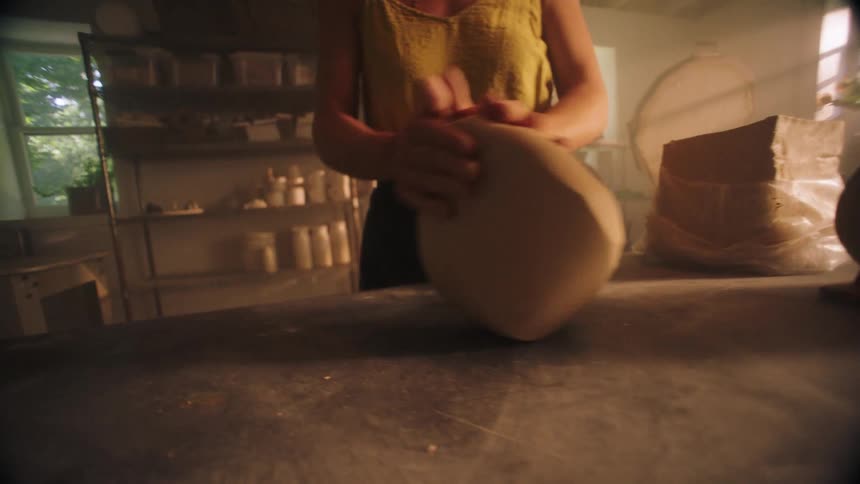 A woman in a yellow top vigorously kneads a large lump of clay on a dusty table in a dimly lit pottery studio
