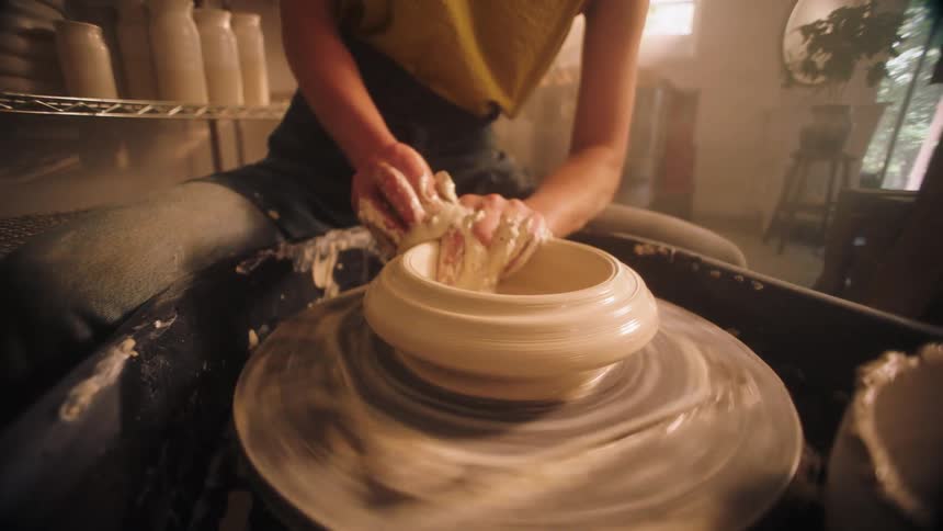 A woman covered in clay skillfully uses her hands and a sponge to refine and open the clay vessel spinning on a pottery wheel in a sunlit studio
