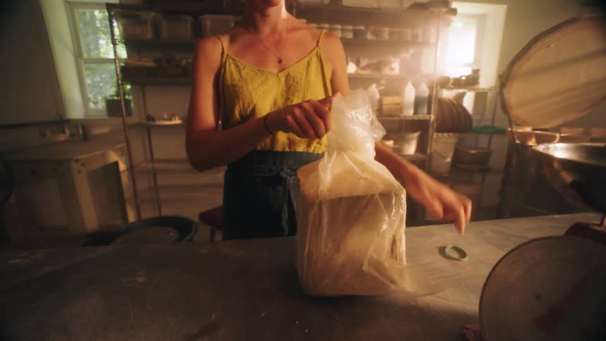 Bathed in the warm glow of a pottery studio a woman stands at her workbench and carefully unwraps a large block of fresh clay peeling away the plastic to prepare the material for sculpting