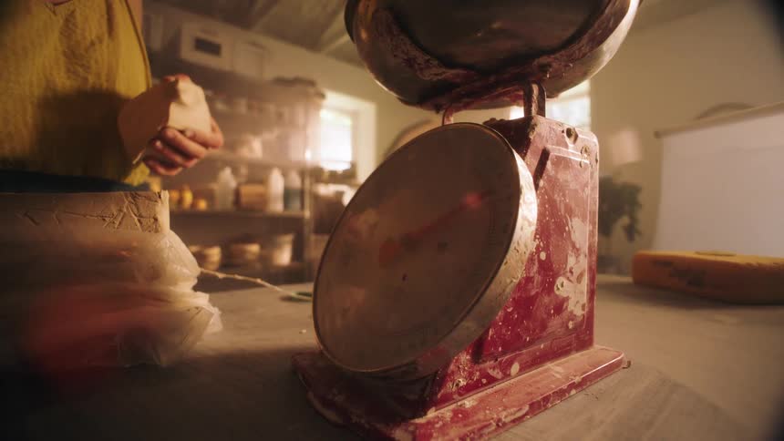 Female potter weighs raw clay on a dirty scale while in a workshop during the day