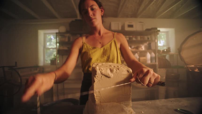 A ceramist in a yellow top unwraps a large block of clay on a table then uses a wire cutter to slice the material in half within a dark warmlit studio