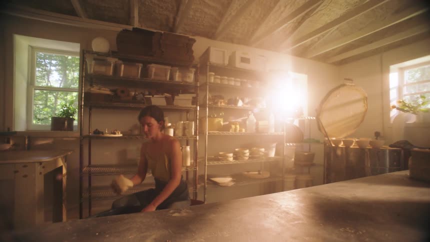 In a sunlit pottery studio a woman kneads a lump of clay on a table before turning and sitting down on a bench near a wall of shelves displaying finished ceramic pieces