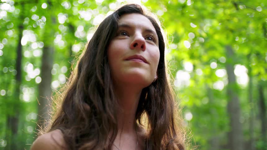 A teenage girl with long brown hair exploring the woods at midday looking around curiously as tall trees and their leaves filter the sunlight