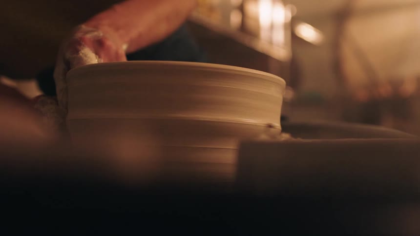 A potters clay covered hands apply pressure to the rim of a spinning clay vessel giving it a final flared shape