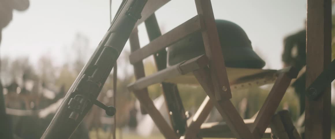 A closeup shot of a boltaction rifle leaning against a wooden folding chair with a dark green military helmet resting on the chair seat
