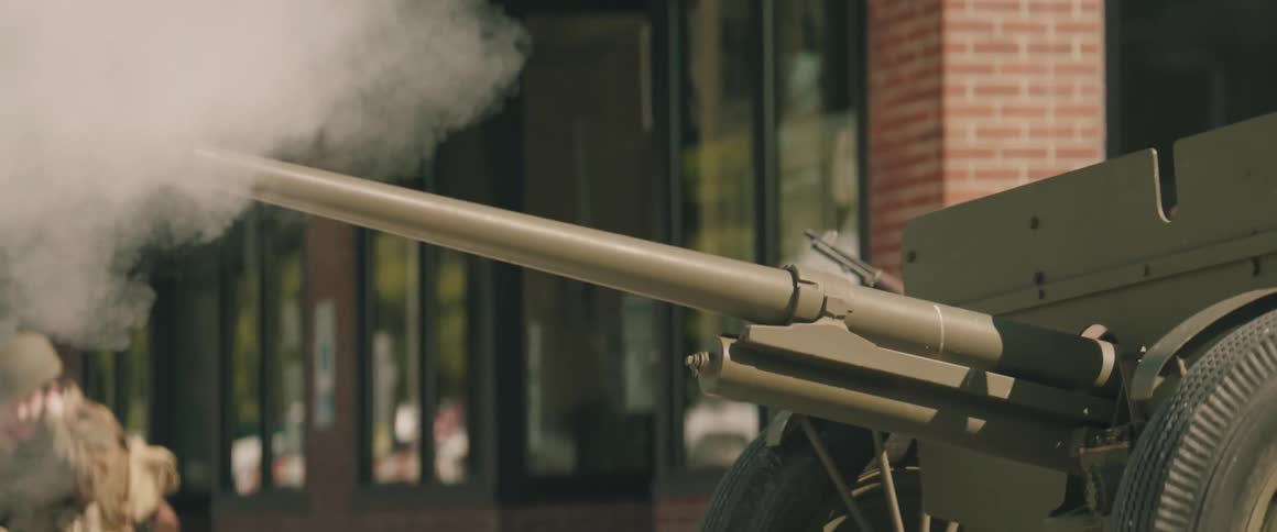 A close view of the muzzle of a military field artillery cannon firing emitting a puff of white smoke in front of a brick building