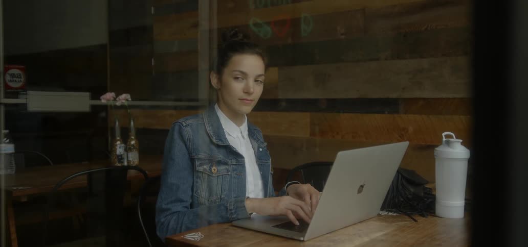 A woman sitting a cafe table is using a laptop and looking out a window