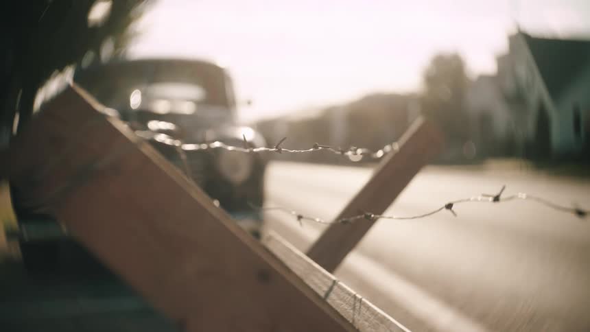 A barbed wire barricade blocks a quiet suburban street with a vintage car and houses softly blurred in the hazy background light