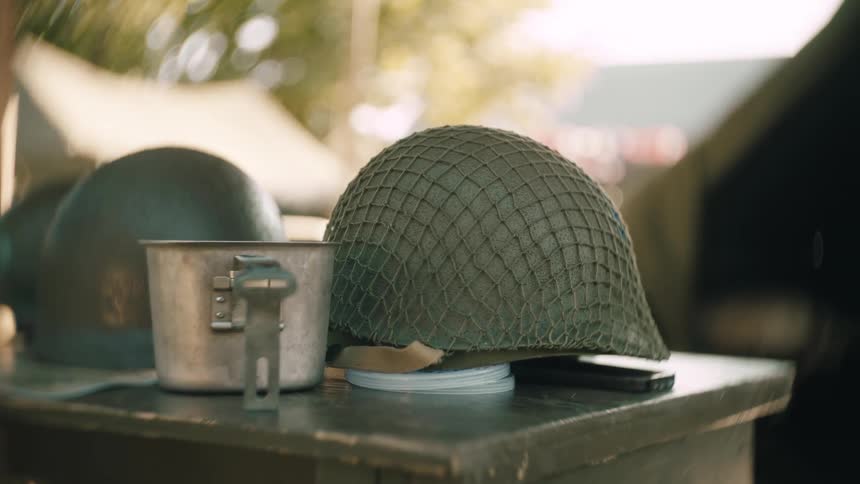 A closeup shot slowly pushes in on a vintage netted army helmet and a metal mess kit sitting on a rustic wooden table at an outdoor camp