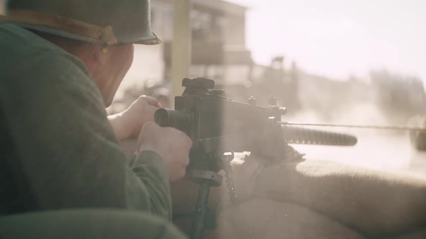 A military personnel crouched behind sandbags on a sunny day wearing a helmet and uniform while firing a machine gun on a sunny day
