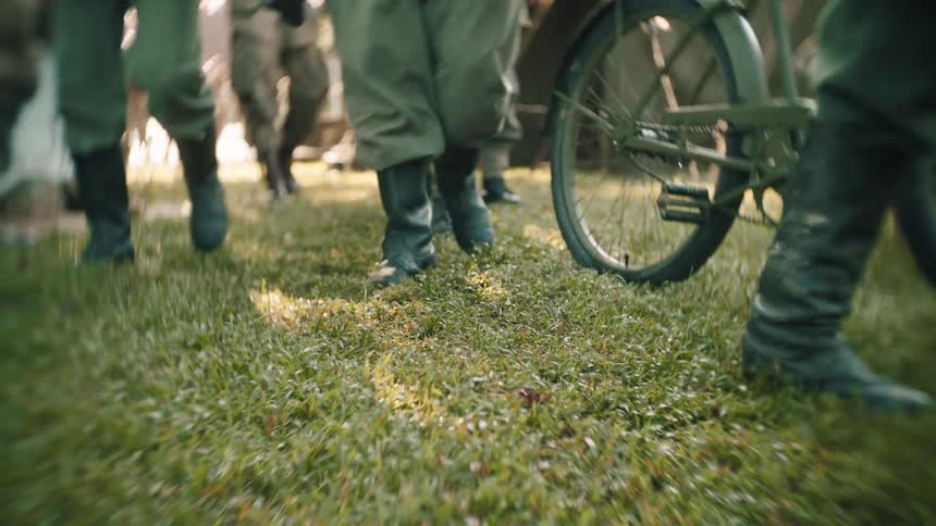 The boots of soldiers in uniform walk across a patch of grass next to a military bicycle